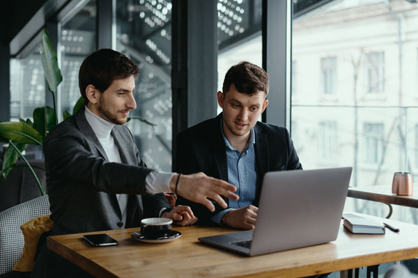 Two businessmen sitting at a table in a modern office setting, discussing something on a laptop screen, with one gesturing while the other listens attentively.