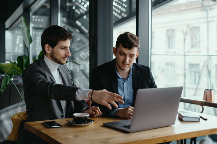 Two businessmen sitting at a table in a modern office setting, discussing something on a laptop screen, with one gesturing while the other listens attentively.