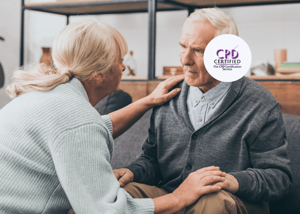 An elderly woman gently supports an elderly man as they sit together at home, sharing a warm and caring conversation.