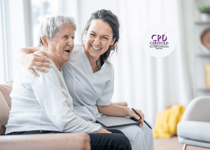 A smiling care worker sits on a sofa with an older woman, both laughing warmly with arms around each other, reflecting trust and positive care relationships.