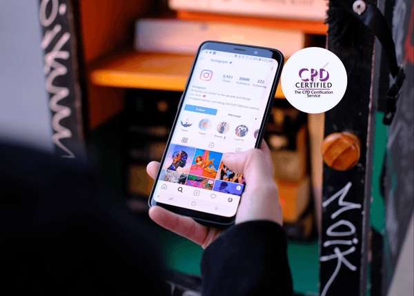 A hand holding a smartphone showing the Instagram profile screen, featuring vibrant image thumbnails.