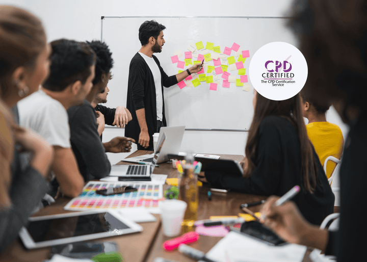Man facilitating a meeting, pointing at sticky notes on a whiteboard, while colleagues seated around a table work with laptops and tablets.
