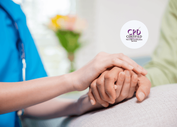 A nurse in a blue uniform gently holds a patient’s hand as they rest in a hospital bed, with a soft-focus bouquet of flowers in the background.
