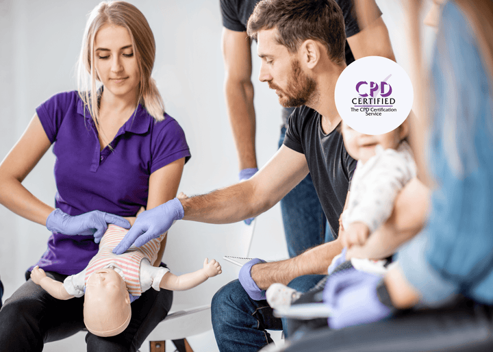 A woman holds an infant manikin while a man wearing purple gloves demonstrates a first aid technique, illustrating paediatric emergency response training.