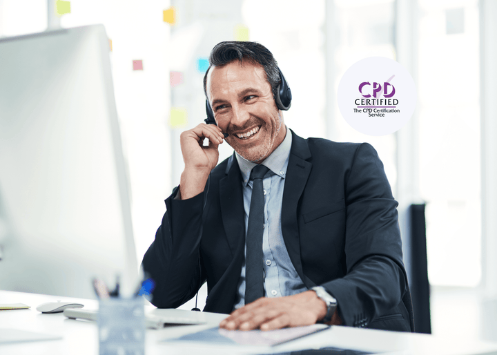 A smiling man in a suit wears a headset while seated at a desk with a computer and notepad, representing professional communication and sales skills in action.