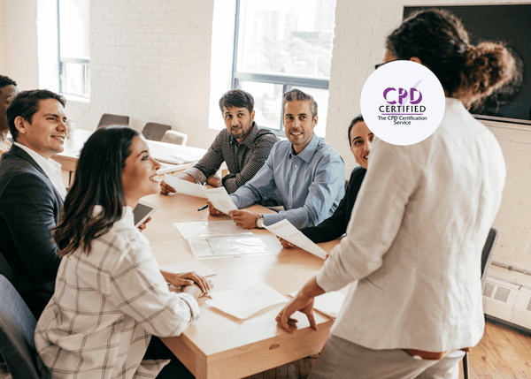 A woman standing at a meeting table, speaking to a group of six smiling colleagues who are holding papers.