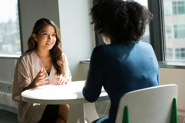 Two women seated at a small round table in an office setting, engaged in a professional conversation near a window with natural light.