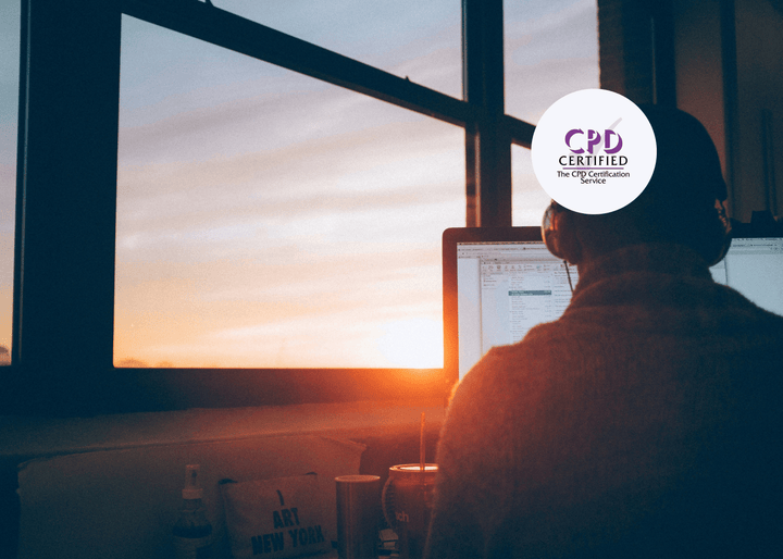 Individual wearing headphones, focused on a desktop computer, seated by a large window with warm sunset light streaming into the workspace.