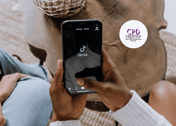 Close-up of hands holding a smartphone showing the TikTok app home screen, with two people sitting at a wooden table in the background.