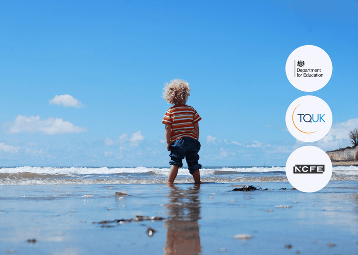 A young child with curly blonde hair wearing a striped shirt stands at the water’s edge on a sandy beach, looking out towards the sea.