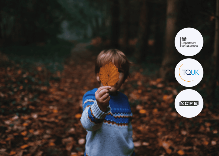 A child stands in a forest holding an autumn leaf up to their face, partially obscuring it from view, creating a playful and seasonal scene.