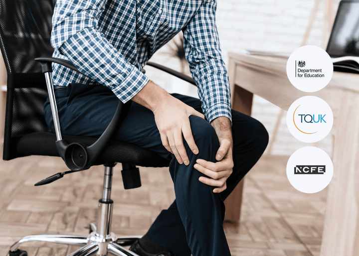 A man in a checked shirt sits in an office chair, holding his knee with both hands and showing visible discomfort, suggesting pain or a health condition.