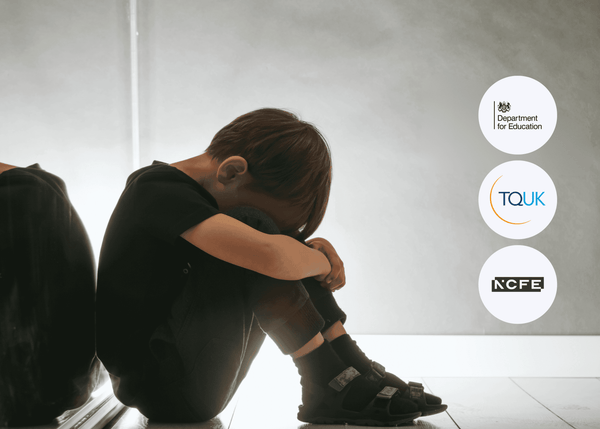 A young child in dark clothing sits on the floor beside a wall, head lowered and arms wrapped tightly around their knees, conveying distress and vulnerability.