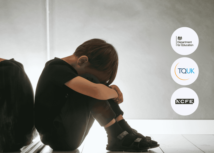 A young child in dark clothing sits on the floor beside a wall, head lowered and arms wrapped tightly around their knees, conveying distress and vulnerability.