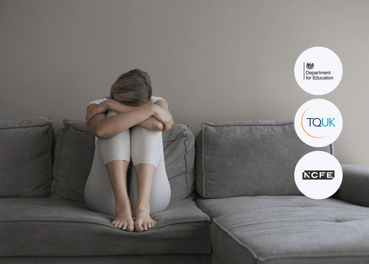 A woman sits curled up on a grey sofa, head resting on folded arms with knees drawn to her chest, in a neutral-toned room, conveying sadness and vulnerability.