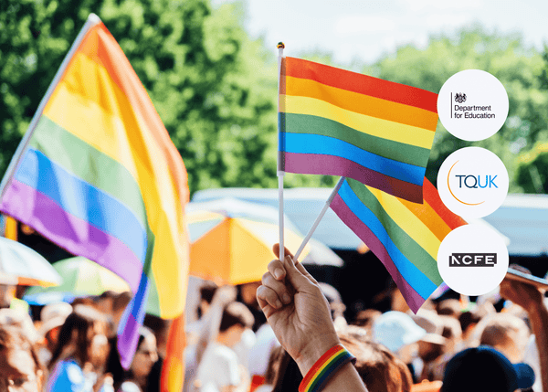 A hand holding a small rainbow flag at an outdoor pride event, with people, umbrellas, and trees in the background, symbolising LGBTQIA+ inclusivity.