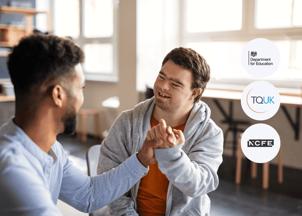 Two people sitting together in a bright room, smiling and clasping hands in a supportive gesture during a casual conversation, symbolising care and inclusion.