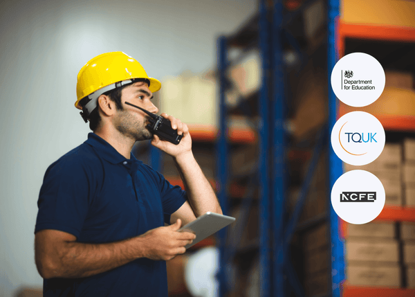 A man wearing a yellow hard hat holds a tablet and speaks into a walkie-talkie while standing inside a warehouse filled with shelves and stacked boxes.