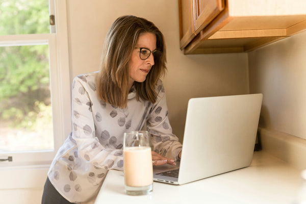 Woman wearing glasses using a laptop at a kitchen counter with a glass of smoothie in front of her, representing learning and working from home.