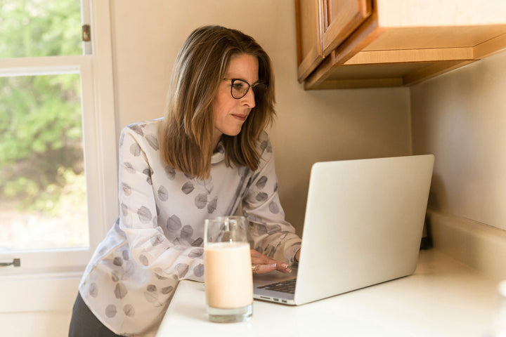 Woman wearing glasses using a laptop at a kitchen counter with a glass of smoothie in front of her, representing learning and working from home.